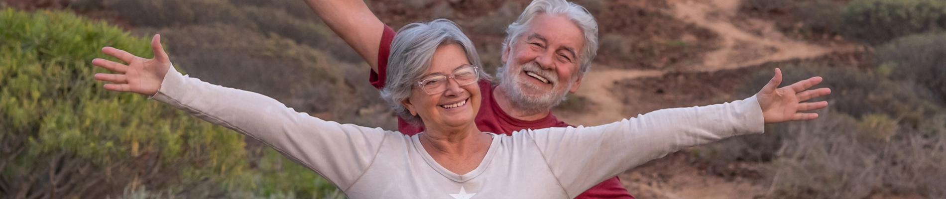 older couple grinning with out streched arms on a hicking trail